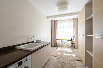 Bright laundry room with sink, counter, washing machine, ironing board by the window, fluffy rug, and cabinets