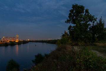 Fototapeta premium Long exposure night photo from Strogino high bank showing Moscow River with poplar silhouette and reflections of illuminated Shchukino residential district under urban glow.