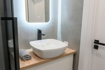 Modern bathroom with a white square sink, black faucet, and illuminated mirror on gray tiles. Neutral tones and sleek design create a clean aesthetic