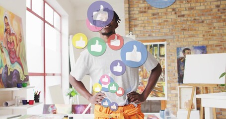 Standing mid adult Black male artist surveying art studio, with floating thumbs-up icons