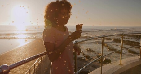 Woman wearing pale pink dress leaning against railing during coastal sunset, holding ice cream cone