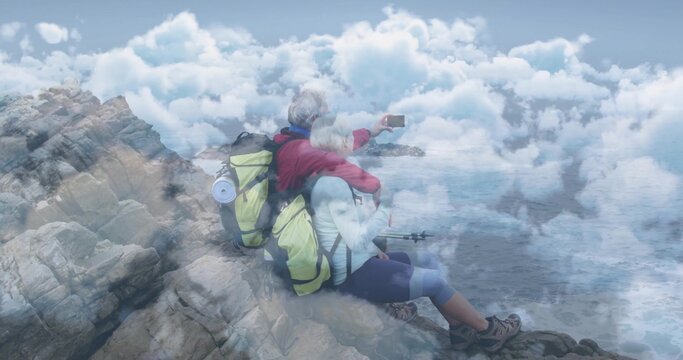 Taking selfie mature hiking couple leaning on coastal edge, with smartphone, backpacks and poles