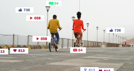 Cycling couple pedaling bikes along seaside path with lamp posts, beach huts, notification overlays