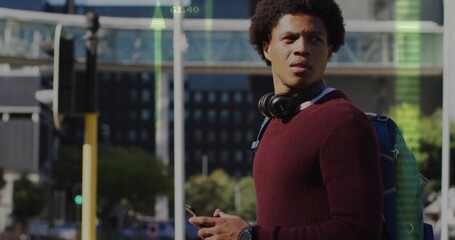 Checking smartphone adult male wearing sweater at urban intersection, with headphones and backpack