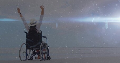 Person in wheelchair raising arms on sandy beach facing sea, with straw hat and wristwatch