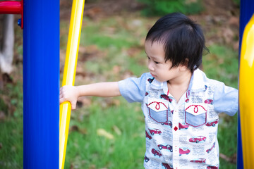 Cute young Asian toddler boy in patterned shirt stands in playground, looking off to the side with...