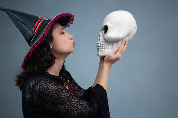 Completely in love witch blowing a kiss to a spooky skull. Ideal for Halloween, romantic horror, bizarre love, witchcraft, and sorcery. Horizontal studio portrait with copy space.