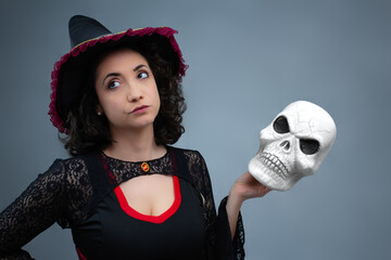 Funny witch rolling her eyes, annoyed, holding a spooky skull in her hand. Ideal for Halloween, creepy comedy, magic, and witchcraft. Horizontal studio portrait with copy space.