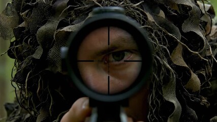 Sniper in camouflage ghillie suit aiming through rifle scope in forest.