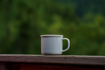 A small white mug with a drink against a background of forest and mountains. Cozy outdoor relaxation