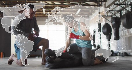 Kneeling trainer and trainee punching torso-shaped heavy bag in boxing gym, with world map overlay