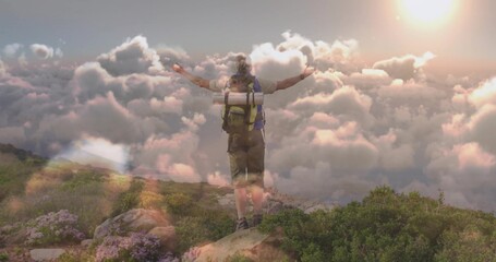 Standing hiker raising arms on rocky ridge above clouds at sunrise, showing backpack gear and boots