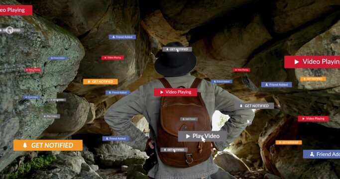 Standing male hiker wearing wide hat viewing rock cave with backpack, camera and digital prompts