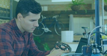 Inspecting engineer examining gear at makerspace bench, with laptop 3D printer bicycle plant boxes