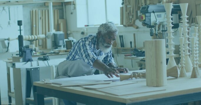 Leaning senior woodworker inspecting paper plans on workshop bench, with prototypes and drill press - Powered by Adobe