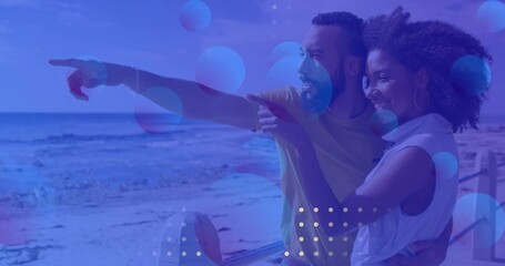 Leaning couple pointing at horizon on metal sandy beach railing, white and yellow tops, copy space