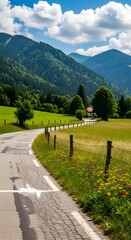 Paved country road winds through green fields and forested mountains under a blue sky with clouds