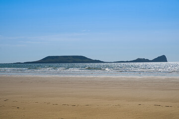 Worm’s Head rises beyond Rhossili’s sandy beach, where sunlit waves roll ashore beneath a bright summer sky.