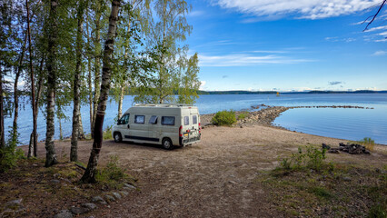 A camper van parked near the serene shores of Lake Saimaa, showcasing the beautiful Finnish landscape with clear skies and gentle waters. Perfect for adventure seekers and nature lovers.