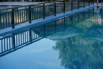 Outdoor swimming pool with clear blue water and reflections of trees and sky. Sun loungers and a black metal fence surround the pool, creating a relaxing holiday and resort atmosphere. Perfect place f