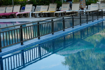 Outdoor swimming pool with clear blue water and reflections of trees and sky. Sun loungers and a black metal fence surround the pool, creating a relaxing holiday and resort atmosphere. Perfect place f
