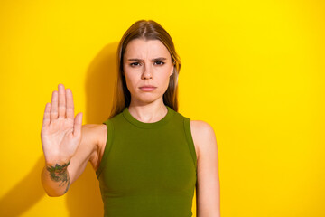 Young blonde woman gesturing stop with hand, wearing green khaki top against vibrant yellow...