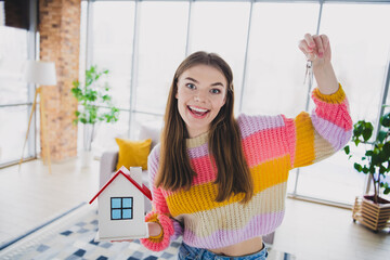 Young woman holding model house and keys happily in cozy living room, expressing excitement and joy of homeownership