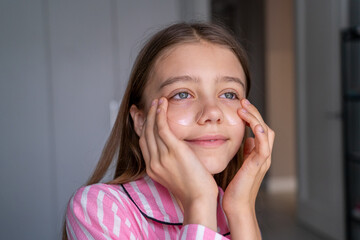 Fototapeta premium Smiling pre-teen girl in pink striped pajamas gently applies hydrating under-eye patches to her skin, enjoying calm morning skincare routine.