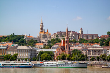 Fototapeta premium Panoramic view of vibrant Budapest cityscape along Danube River, showcasing iconic Matthias Church, historic spires, and diverse architecture, with multiple boats docked on sunny day.
