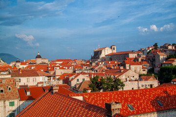 Panoramic high-angle view of picturesque old city, highlighting vast expanse of traditional terracotta tiled roofs, historic stone architecture, prominent church domes, and vibrant blue sky.