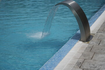 Water jet fountain flowing into a clear blue swimming pool. Modern spa or hotel poolside scene, relaxing and refreshing atmosphere. Ideal for travel, leisure, wellness, or architecture themes