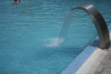 Water jet fountain flowing into a clear blue swimming pool. Modern spa or hotel poolside scene, relaxing and refreshing atmosphere. Ideal for travel, leisure, wellness, or architecture themes