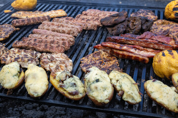 variety of grilled meats and roasted potatoes on a barbecue. The image captures the textures and colors of the food, including grilled sausages, pork ribs, chicken, alongside cheesy roasted potatoes