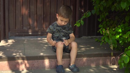 The child looks closely at his scraped knee. He gently touches the scab with his finger. He is sitting on a wooden porch on a hot summer day. He is wearing a t-shirt, shorts and slippers.