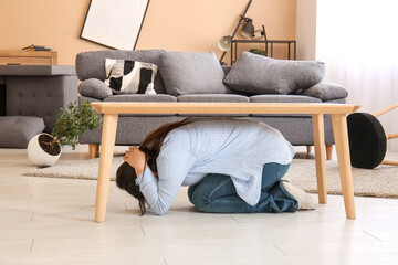 Young woman hiding under table in damaged living room after earthquake