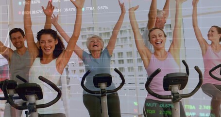 Spin riders pedaling stationary bikes in gym wear raising arms in fitness studio, with stock charts