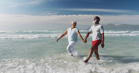 Strolling retirees in beachwear through surf on sandy shore with ocean waves and distant hills
