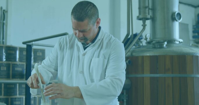 Stirring technician in white lab coat mixing clear liquid in flask at distillery, gin boxes, tank