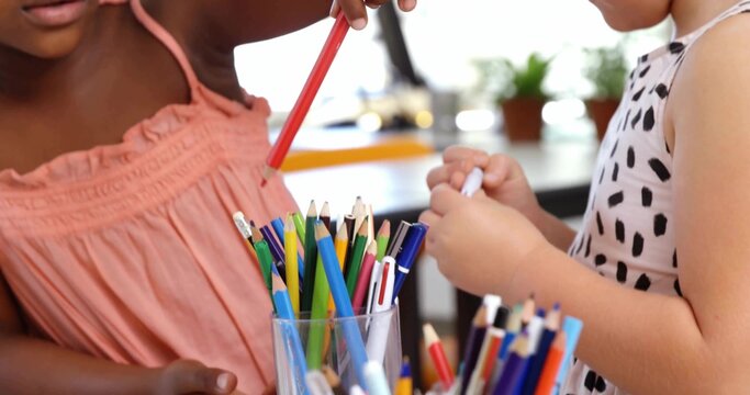 Two girls wearing peach-pink top and spotted dress grabbing from container at desk, colored pencils