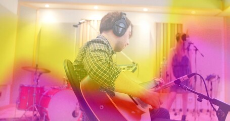 Recording musician strumming electric guitar in recording studio with drum kit left, vocalist right