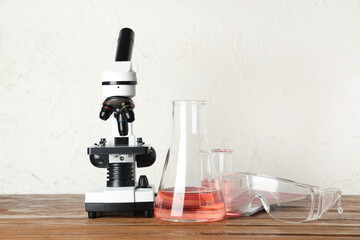 Modern microscope with chemical flasks and protective glasses on wooden table against white grunge background