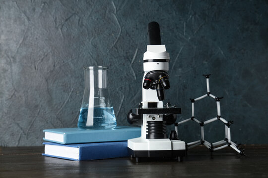 Modern microscope with chemical flask, books and molecular model on wooden table against grey grunge background
