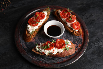 Plate of tasty tomato bruschetta with ricotta and bowl of sauce on black background