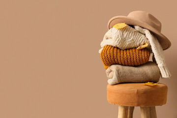 Stack of stylish clothes, hat and autumn leaves on stool against beige background