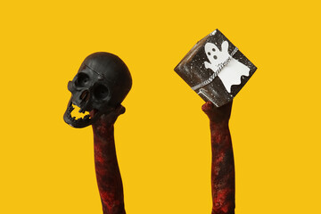Hands of zombie with gift box and skull on yellow background. Halloween celebration