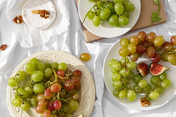 Plates with sweet ripe grapes, walnuts and camembert cheese on white fabric background