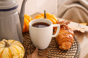 Teapot with cup of hot beverage, pumpkins, croissant and autumn leaves on table in room