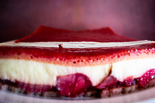Slice of layered sponge cake with creamy filling and glossy red berry jelly, topped with delicate white chocolate strands. Vibrant colors and rich textures create an elegant, festive dessert scene. - Powered by Adobe