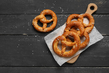 Cutting board with tasty pretzels on black wooden background, closeup
