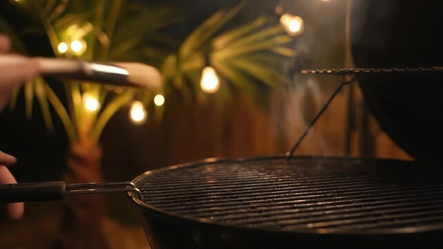 Hand applying marinade with brush to grill grates in outdoor setting, illuminated by warm lights, showcasing the preparation process for a delicious barbecue experience